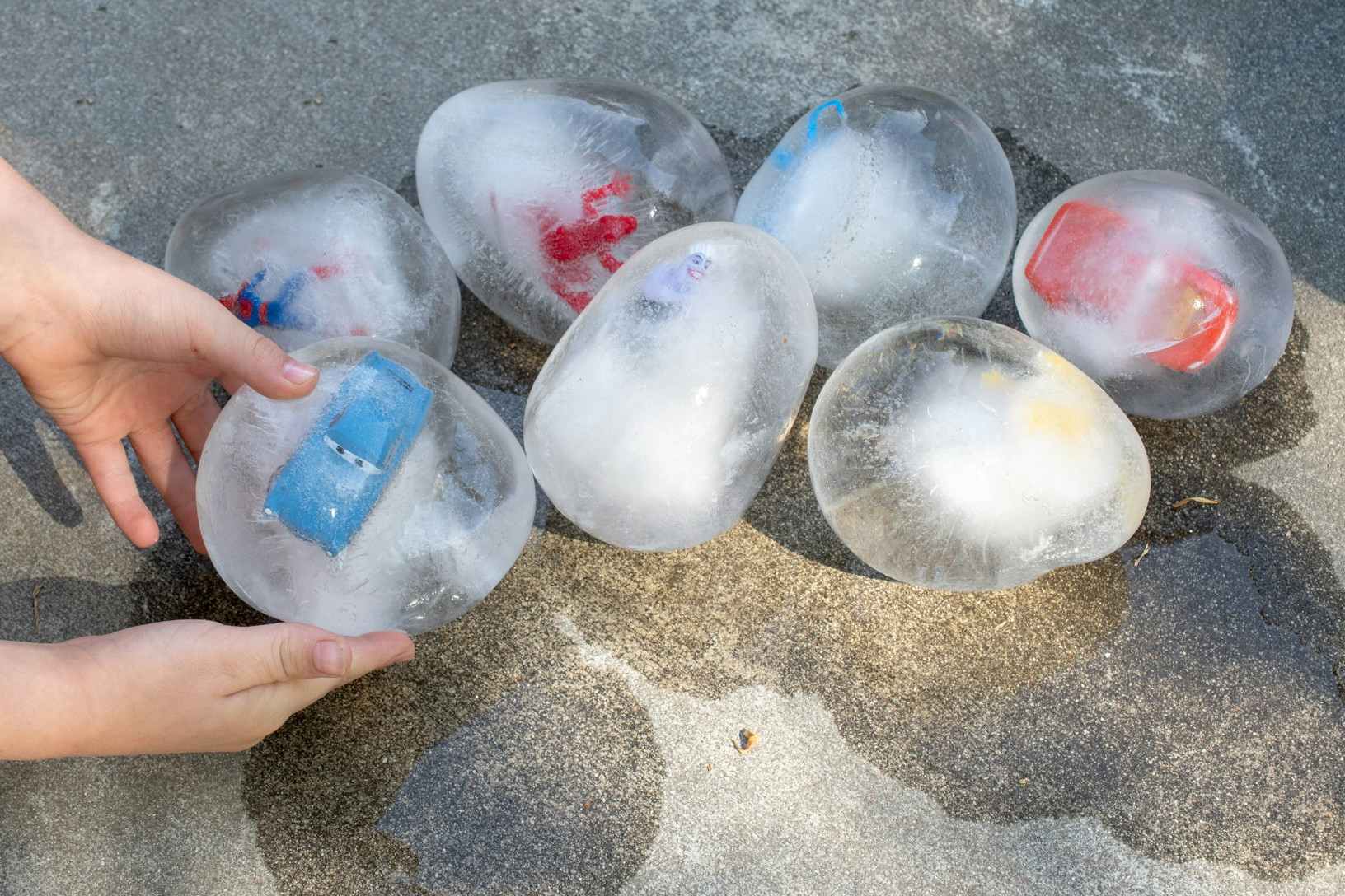 A person's hands picking up a DIY ice egg with a child's toy frozen inside that is sitting on the ground next to similar ice eggs.