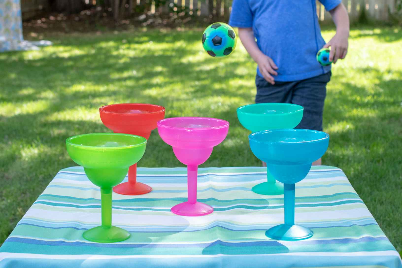Five colorful, plastic margarita cups sitting on a table in a backyard while a child is throwing balls trying to get one in a cup.