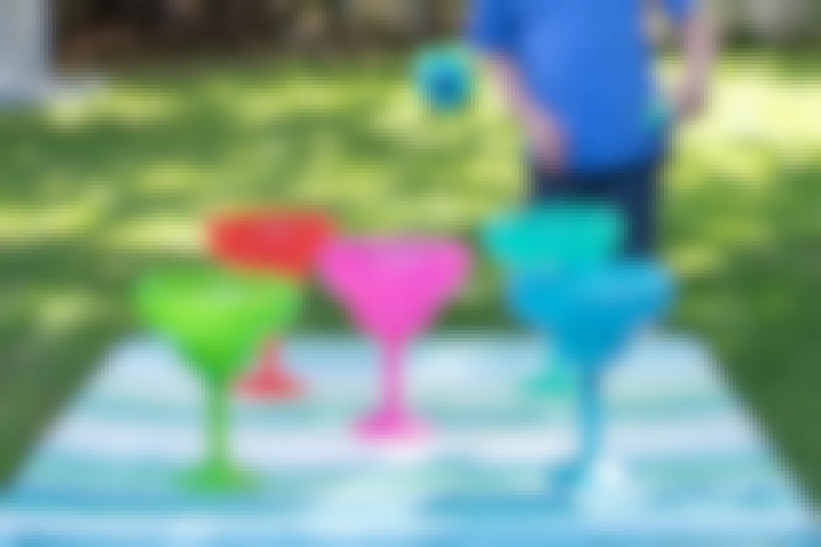 Five colorful, plastic margarita cups sitting on a table in a backyard while a child is throwing balls trying to get one in a cup.