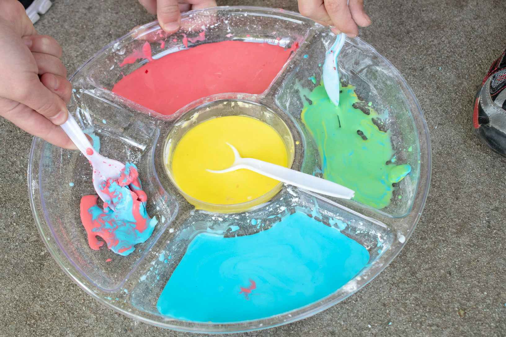 A person's hand using a spoon to mix paint in a repurposed vegetable tray, each compartment with a different color of paint.