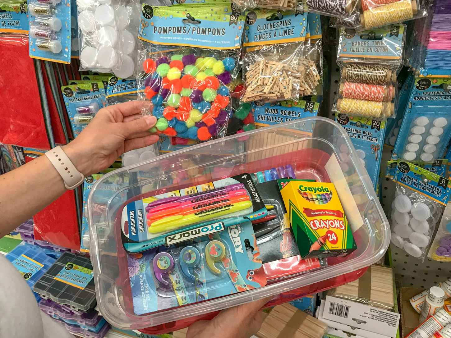 A person's hands holding a tupperware tub filled with art supplies next to a wall of art and craft supplies at a store.