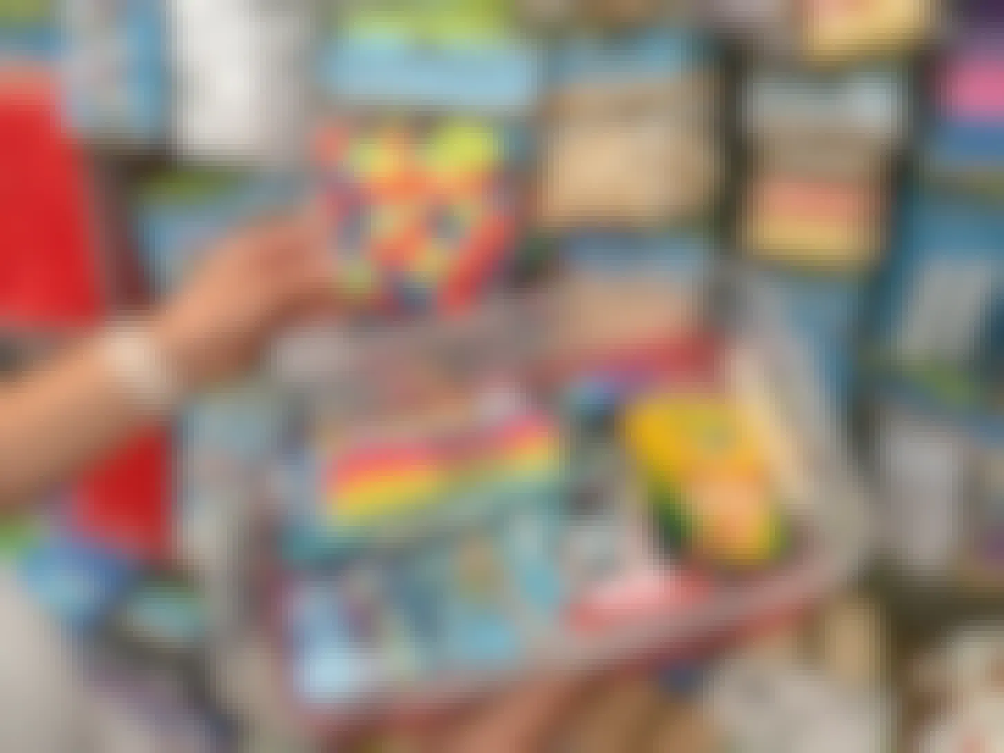 A person's hands holding a tupperware tub filled with art supplies next to a wall of art and craft supplies at a store.