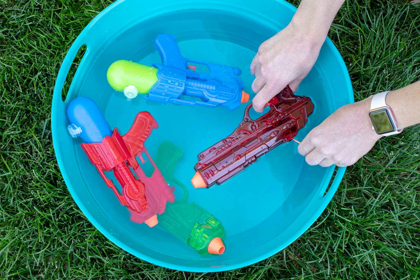 A person's hands reaching to take a water gun from a bucket filled with water and two more water guns.