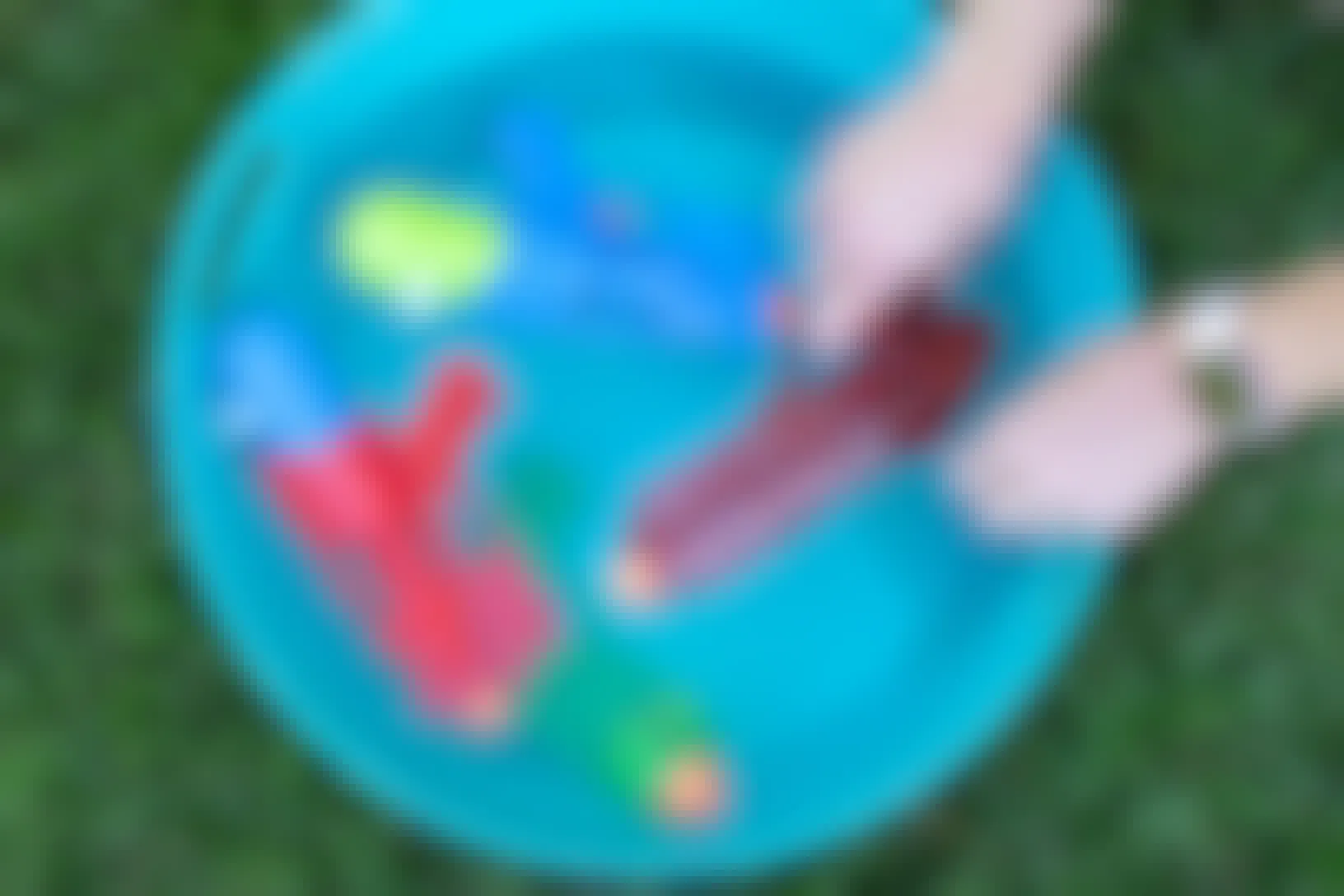 A person's hands reaching to take a water gun from a bucket filled with water and two more water guns.