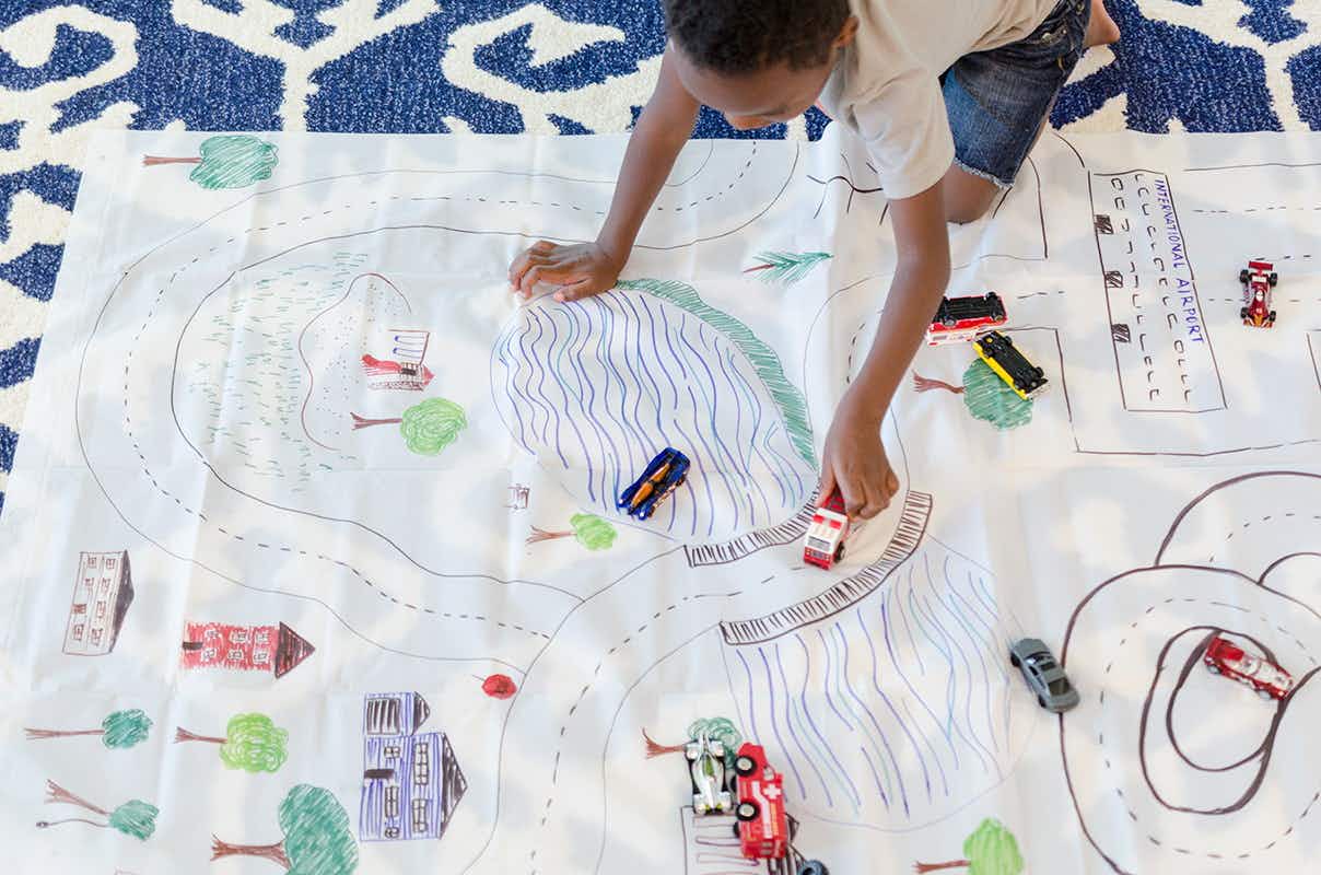 A child kneeling down, playing with a toy car on a shower curtain that has been painted to look like a town with roads. 