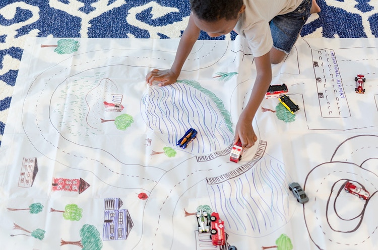 A child kneeling down, playing with a toy car on a shower curtain that has been painted to look like a town with roads. 