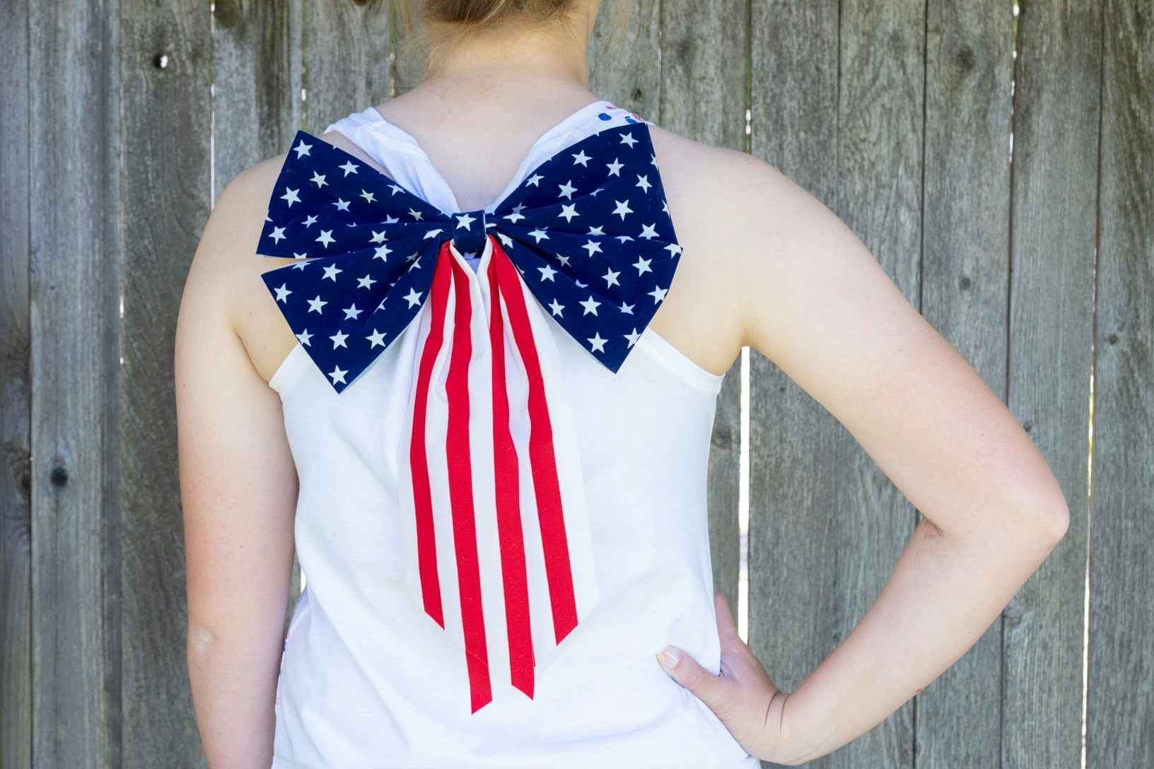 A decorative American flag bow attached to the back of a person's white tank top, standing facing away toward a fence.