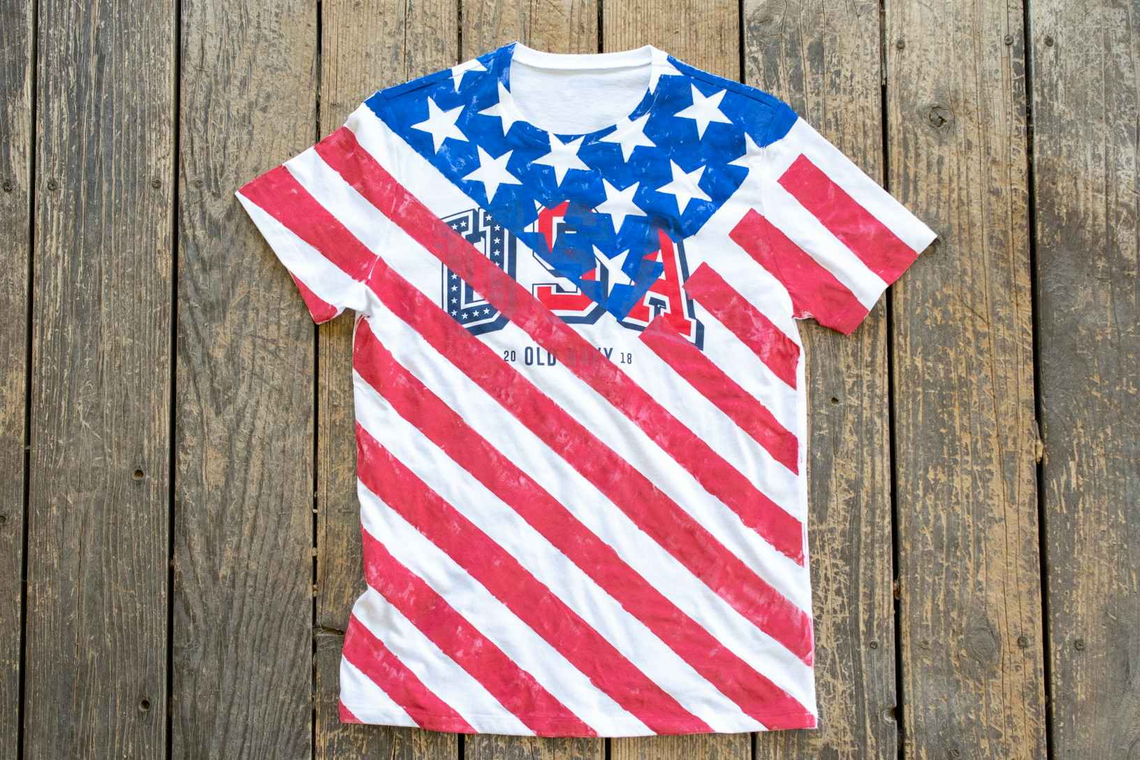 A white Old Navy USA T-shirt painted to look like the American flag laying on a wood deck.
