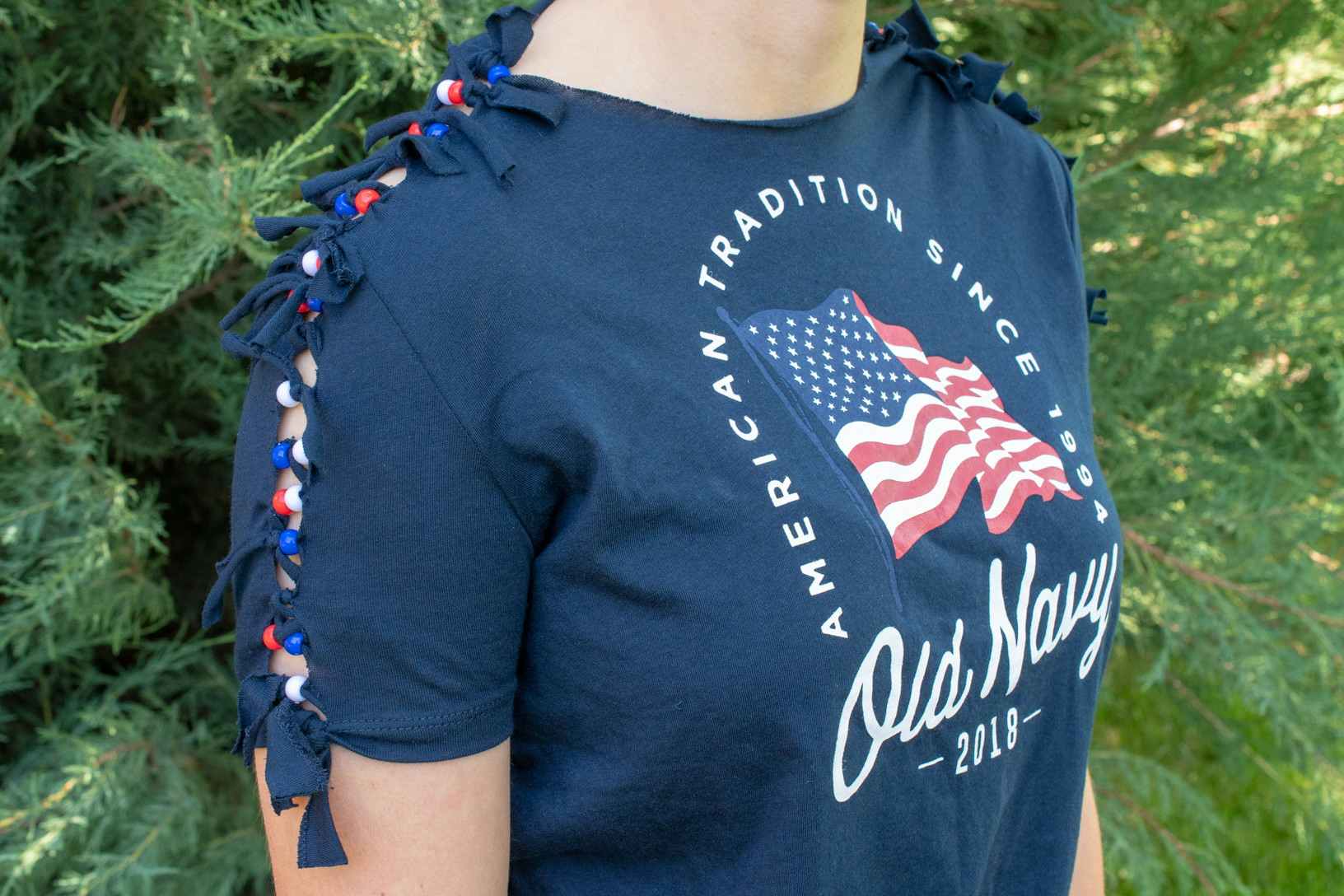 A person wearing an Old Navy Fourth of July t-shirt that has had the sleeves cut and then re-tied with red, white, and blue beads.