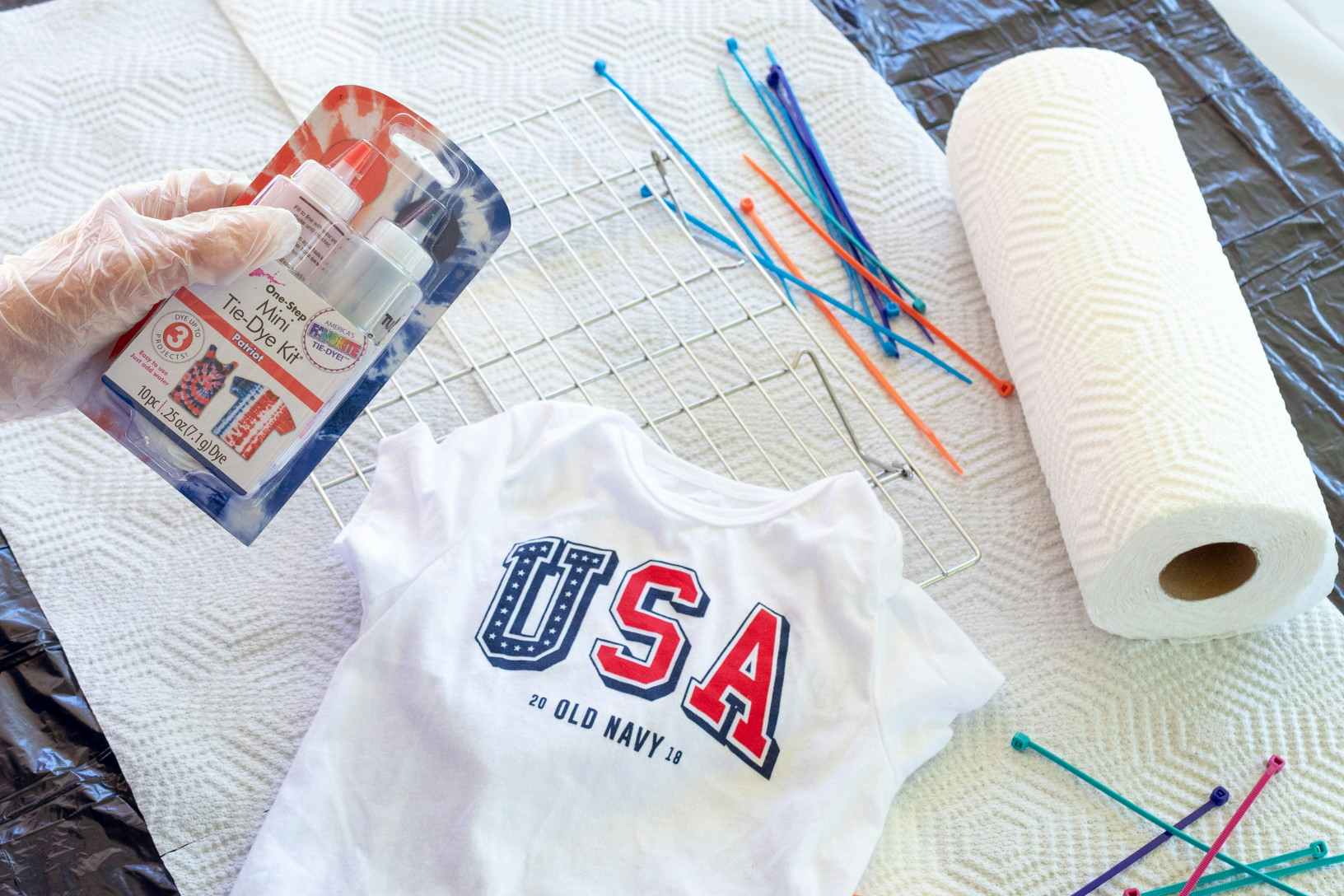A person's gloved hand holding a tie-dye kit above a plain white Old Navy USA baby onsie, some zip ties, and a roll of paper towel sitting on a table.