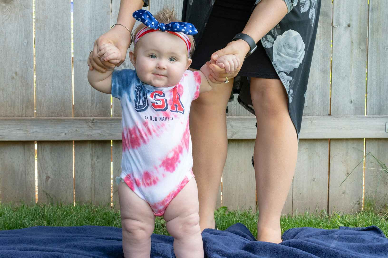 A mother helping her baby stand up to show off the tie dye USA onsie and American flag bandana the baby is wearing.