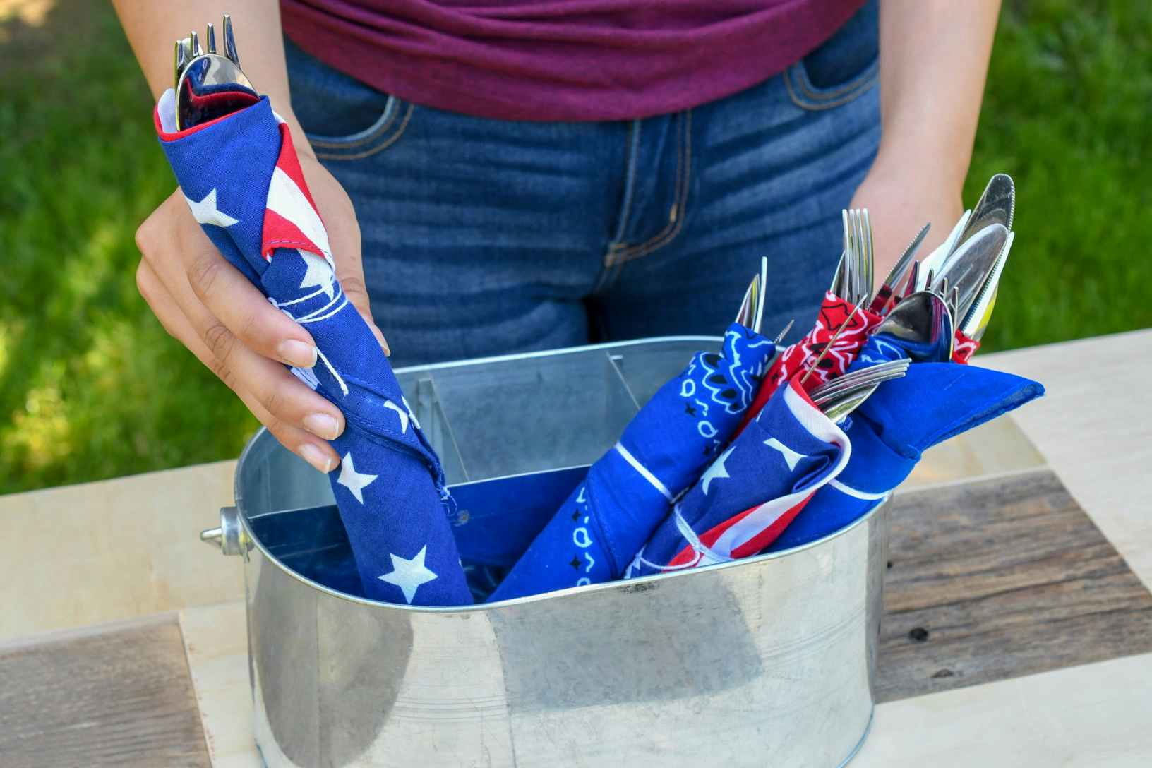 A person putting a set of silverware wrapped in a red, white, and blue bandana into a container with similarly wrapped silverware sets.