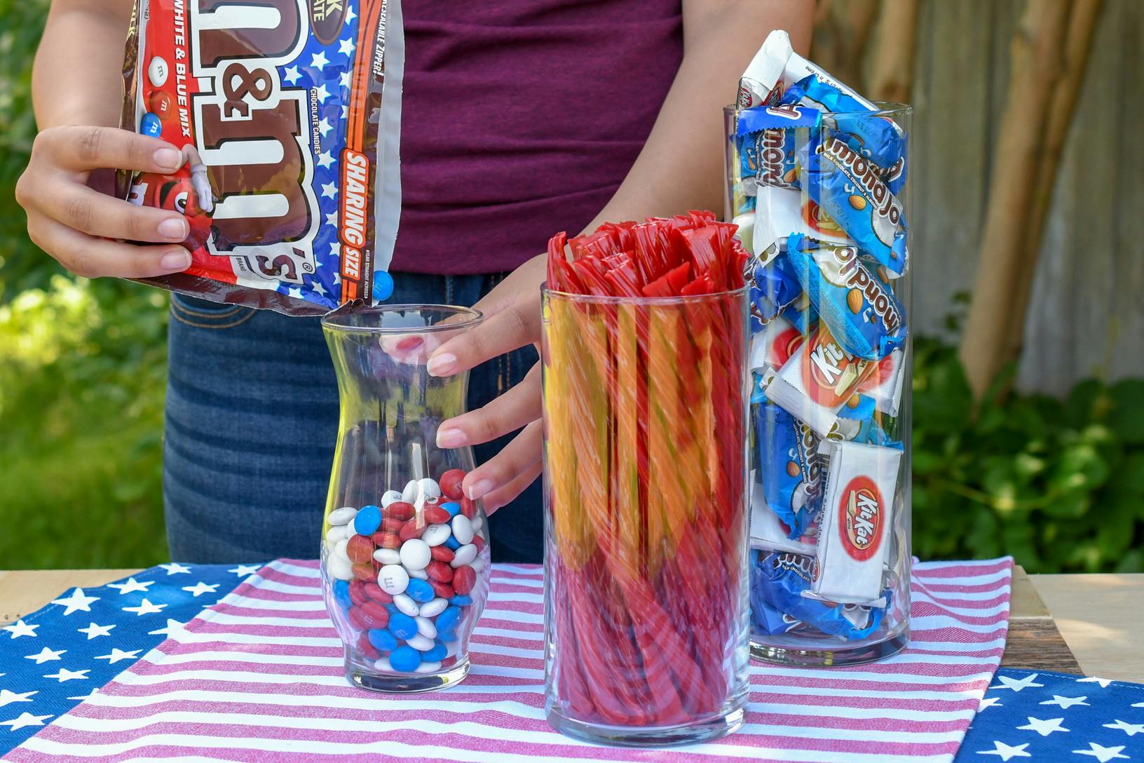A person pouring red, white, and blue M&Ms into a glass vase next to two more glass containers filled with licorice and mini candy bars.