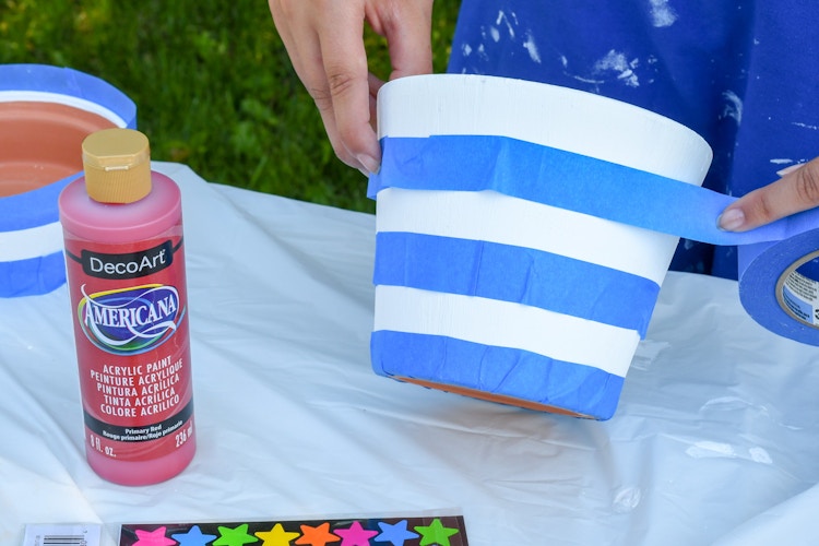 A person using painters tape to put stripes on a terracotta pot on a table next to a bottle of red acrylic paint.