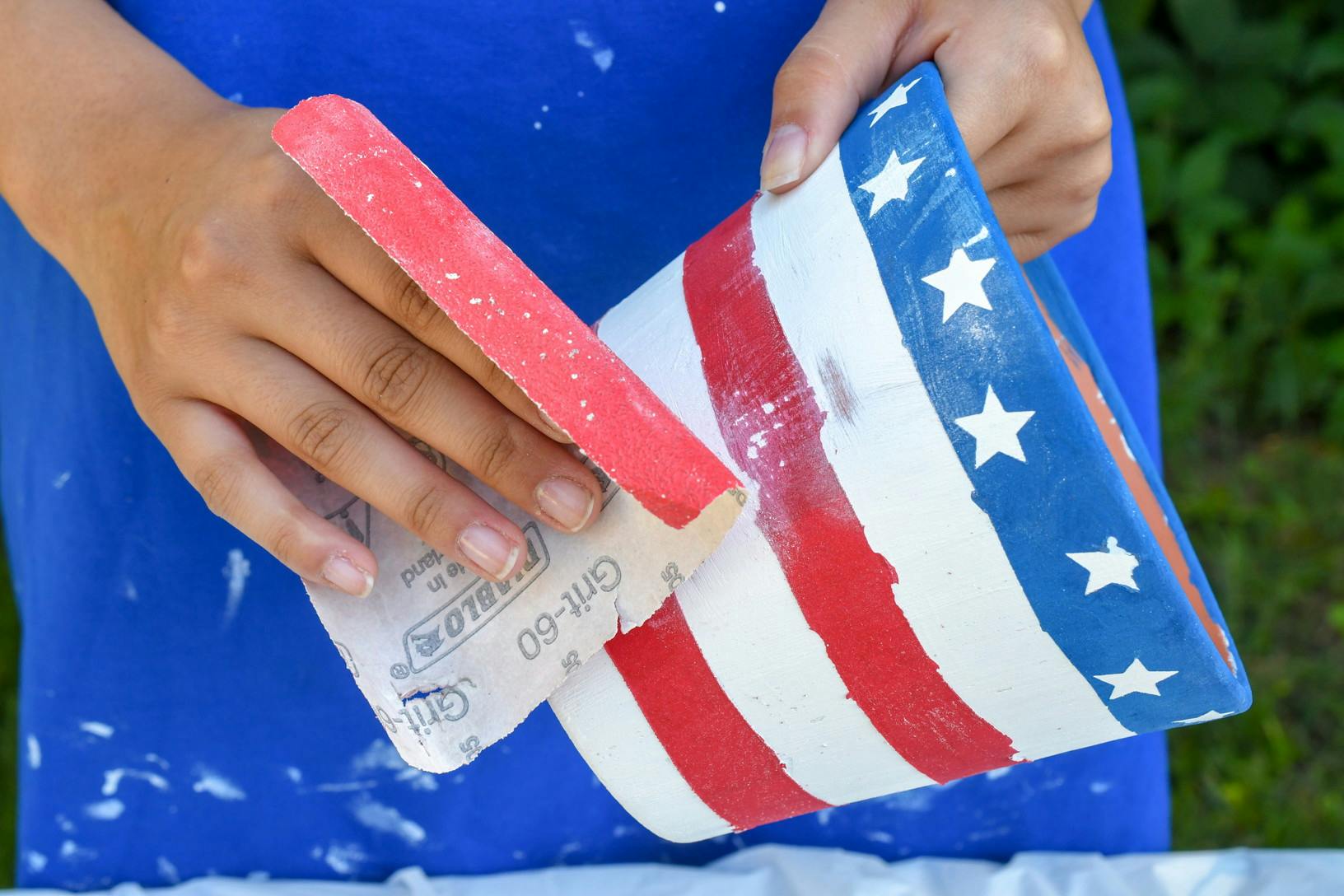 A person lightly sanding a terracotta pot that has been painted to look like an American flag.