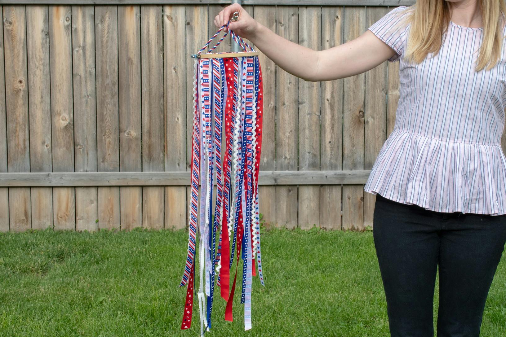 A person standing in a backyard, holding up a DIY windsock made of ribbon and an embroidery hoop.