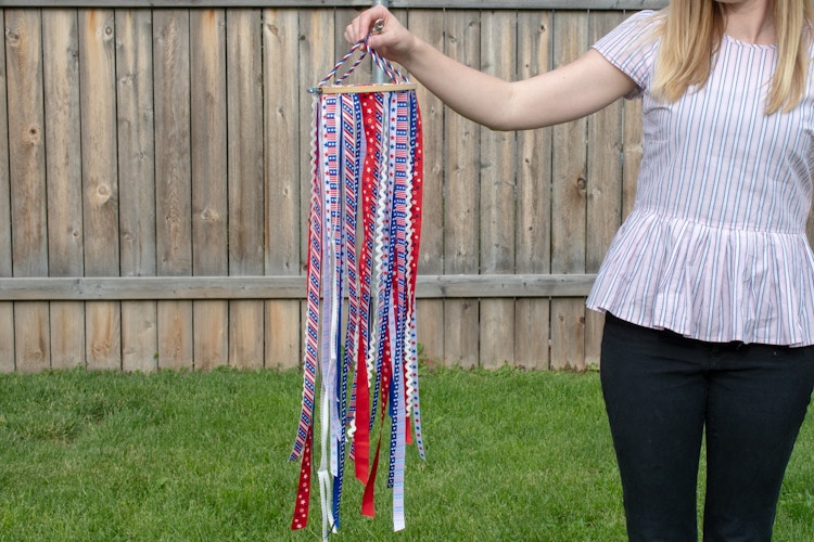 A person standing in a backyard, holding up a DIY windsock made of ribbon and an embroidery hoop.