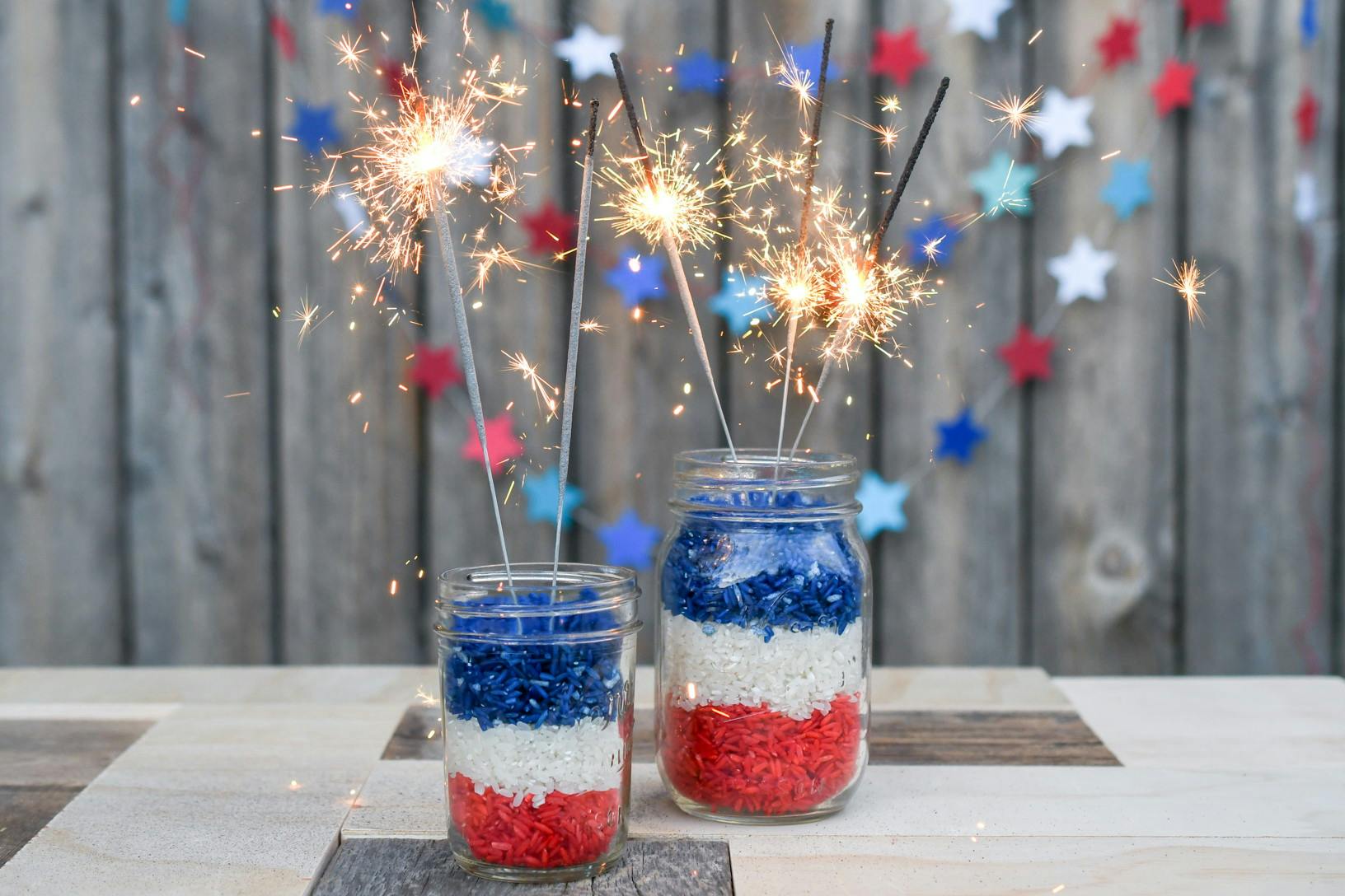 Lit sparklers sticking out of mason jars filled with red, white, and blue rice that are sitting on a table outside.