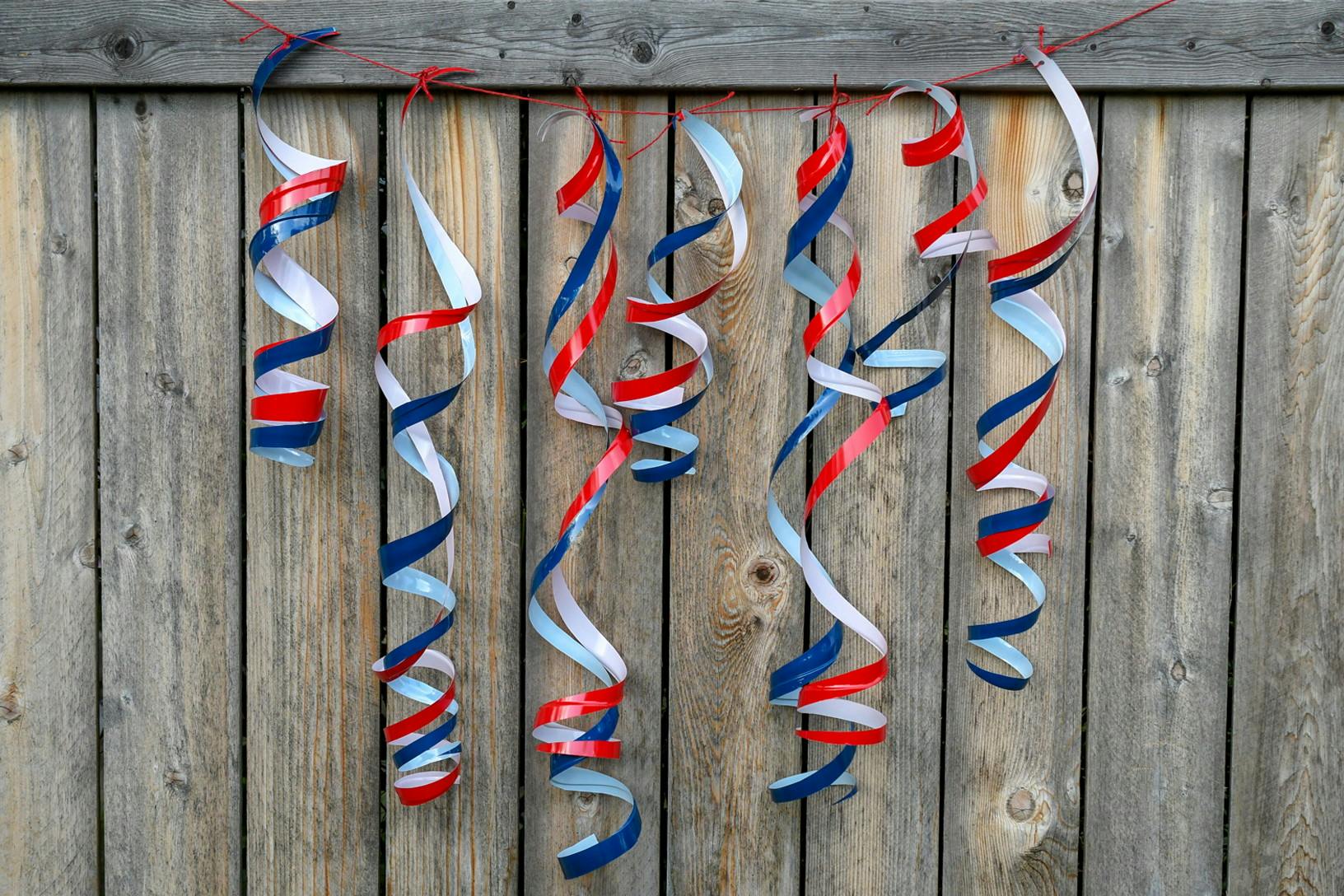 Twirly streamers hung together as a banner made from spiral-cut red and blue plastic cups, hanging on a wooden fence.