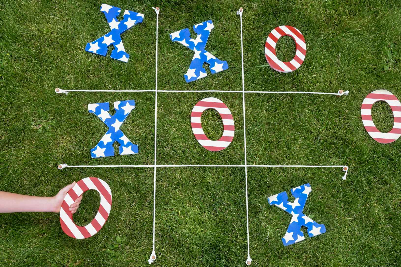 A person placing a red-and-white-striped O in a space below a blue X with white stars on a lawn tic-tac-toe game.