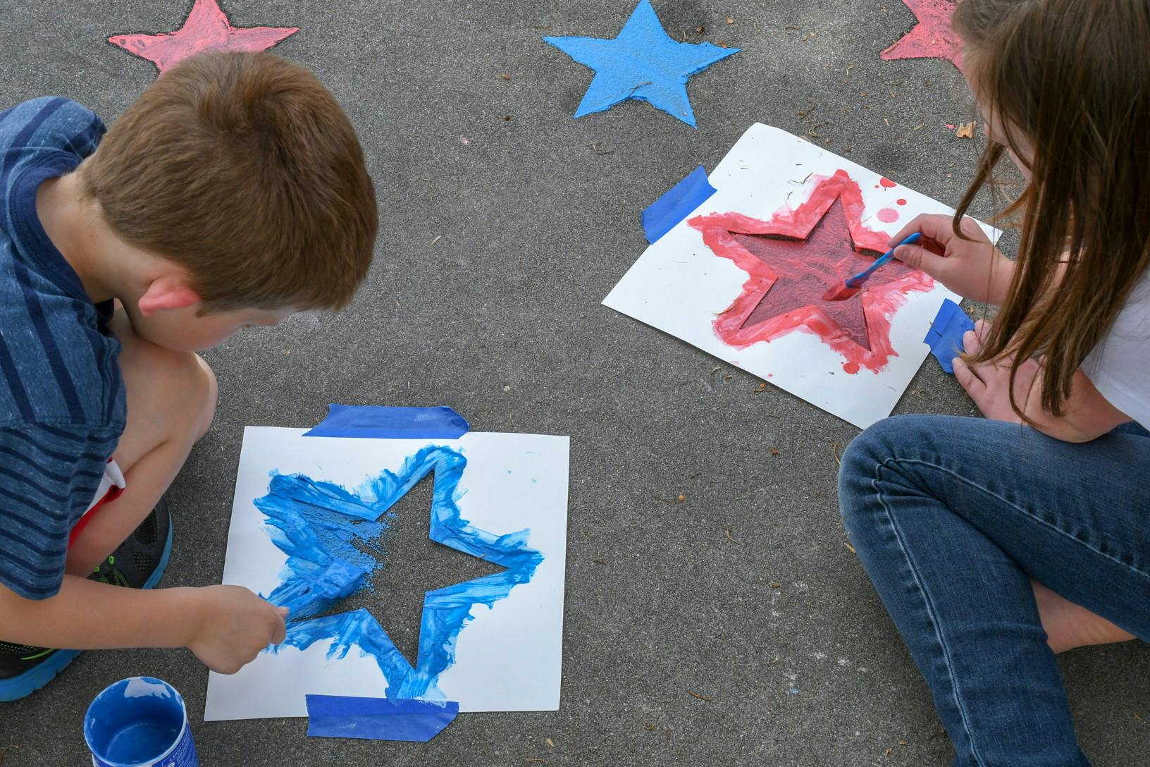 Two children using star-shaped stencils and sidewalk chalk paint to paint red and blue stars onto a driveway.