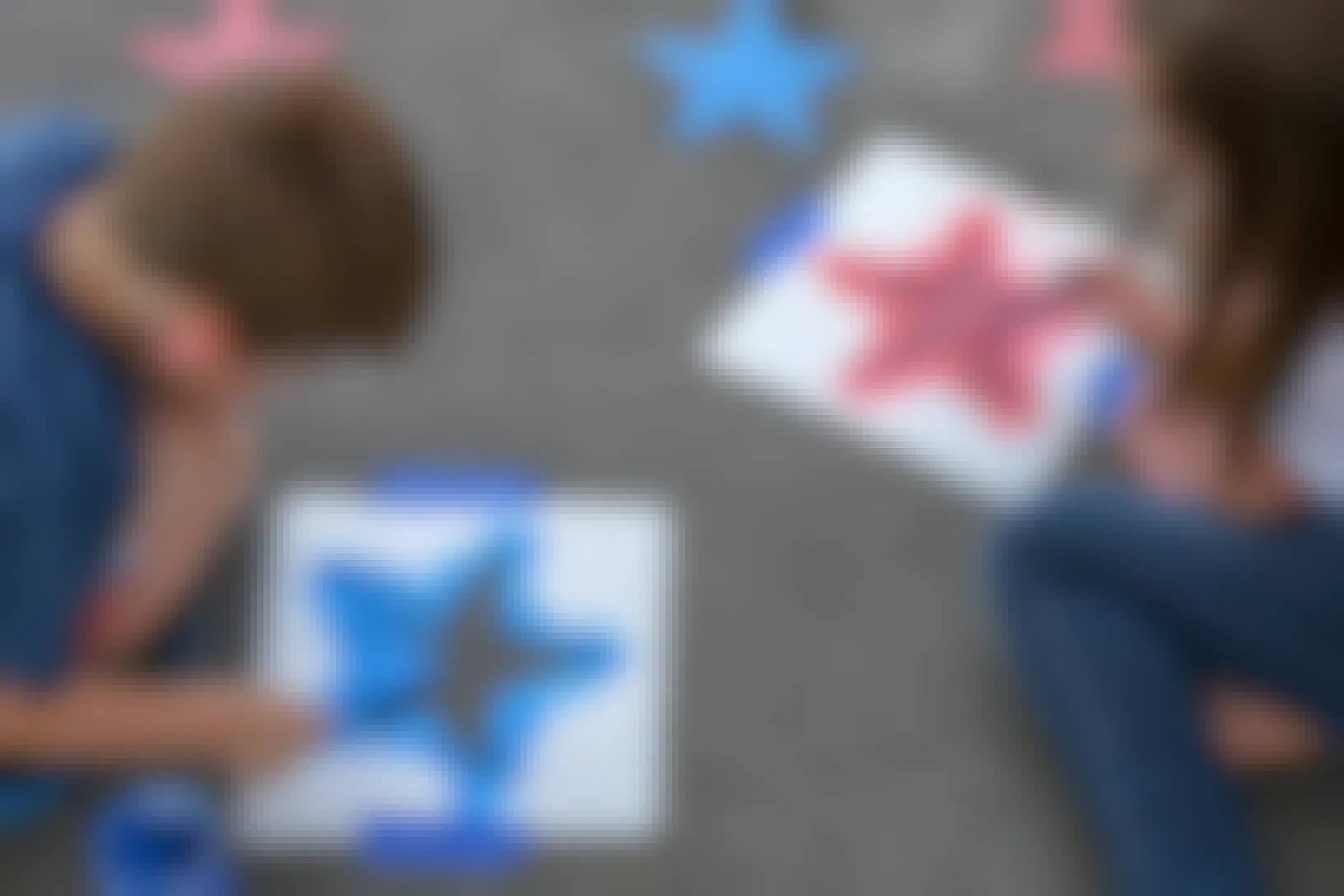 Two children using star-shaped stencils and sidewalk chalk paint to paint red and blue stars onto a driveway.