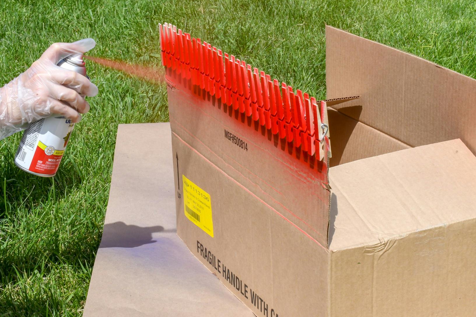 A person spraying red pray paint onto clothespins that are clipped onto the flap of a cardboard box sitting in the backyard.