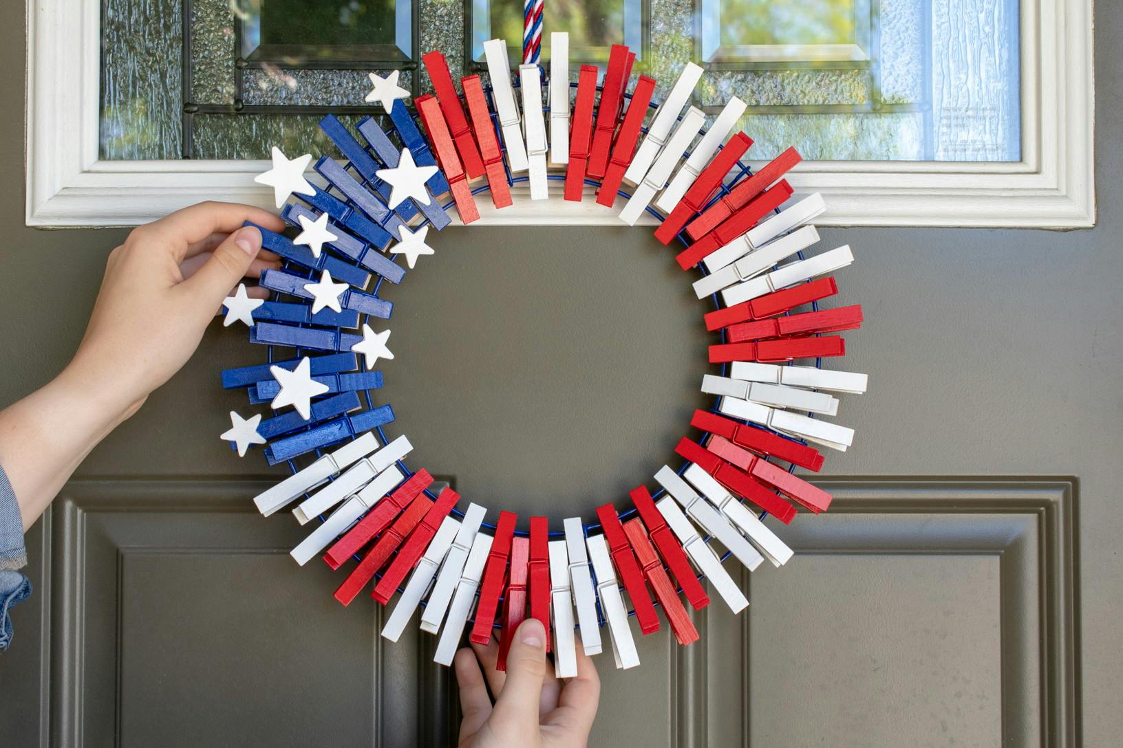 A person hanging a wreath made of red, white, and blue clothespins on their front door.
