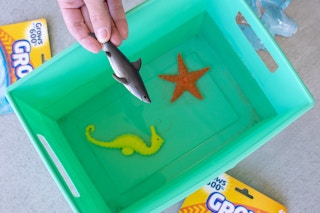 A person's hand holding a toy shark above a square bucket filled with water, with a seahorse toy and starfish toy sitting at the bottom.