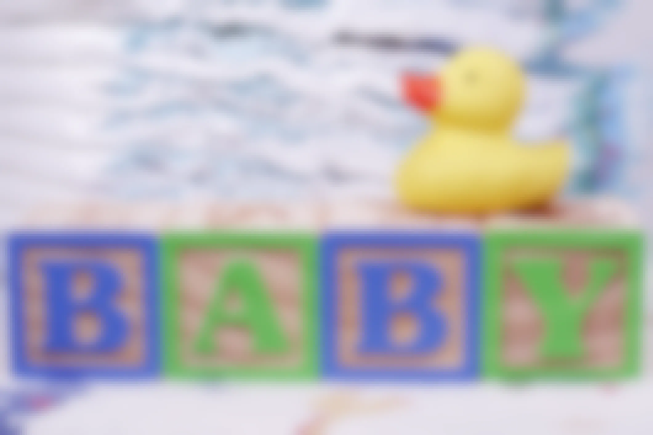 Some baby blocks arranged to spell the word "BABY" with a rubber duck on top and stacks of diapers in the background.