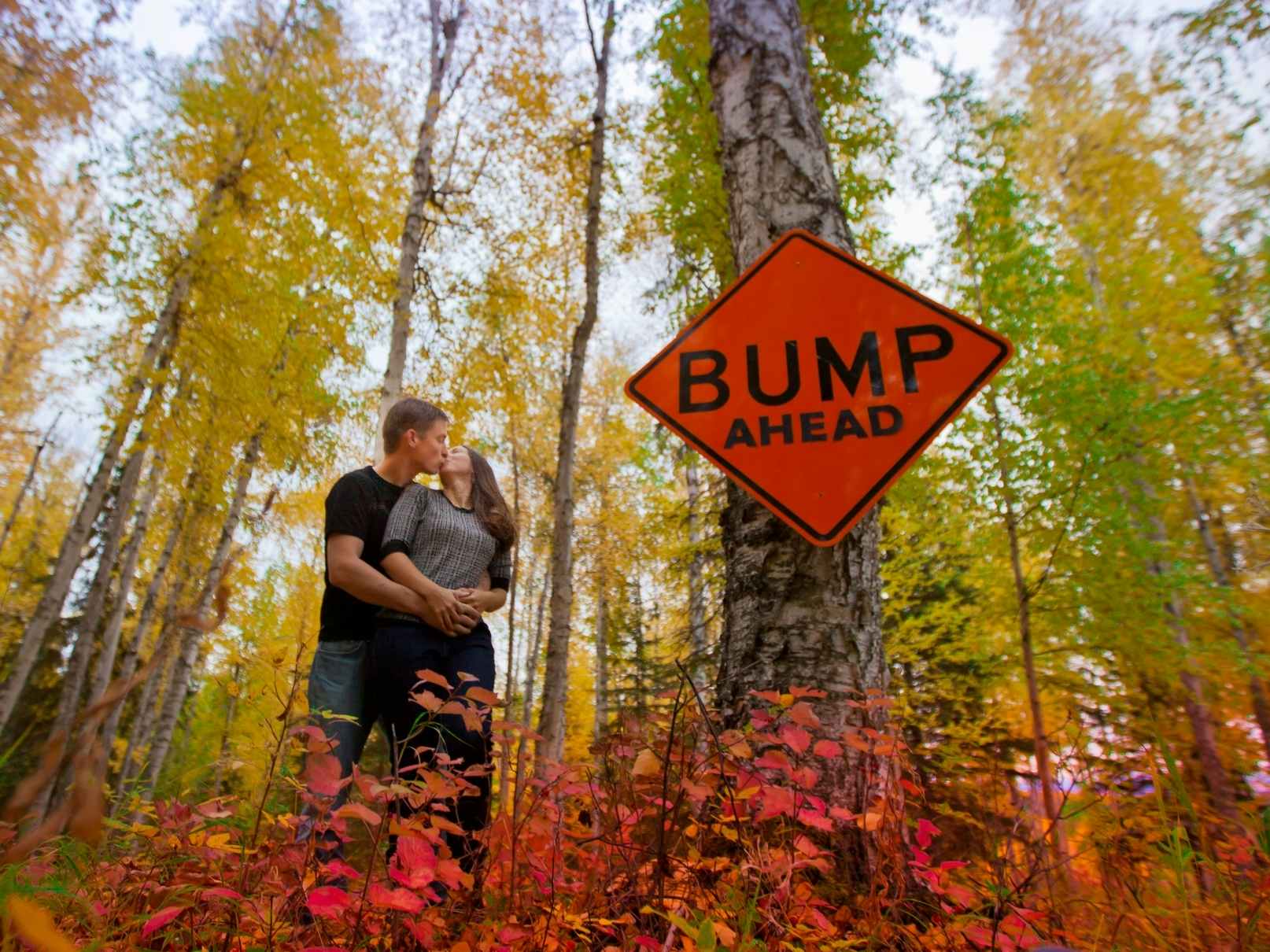A couple kissing in some woods with a sign that reads "Bump Ahead" in front of them.