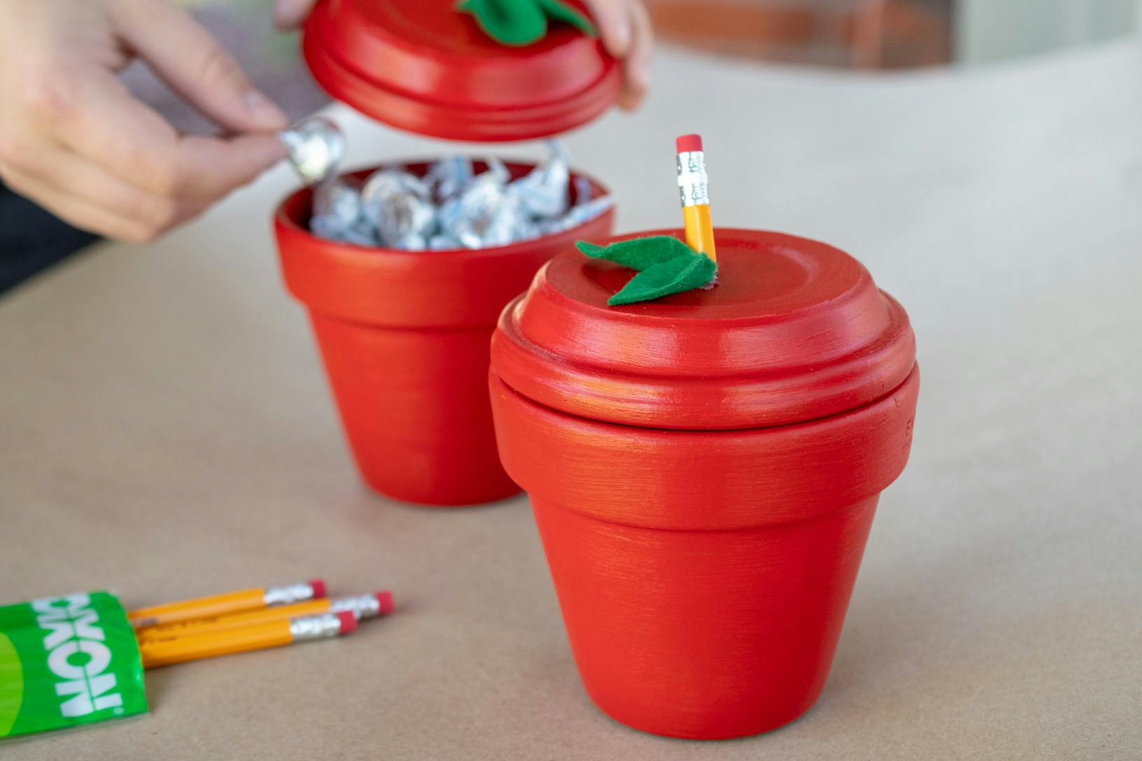 Person grabbing a small candy out of one if two small terra cotta pots painted red with a leaf and pencil end glued to the lids to resemble an apple.