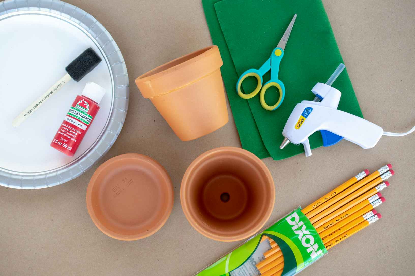 paper plates, paint, terracotta pots, hot glue, scissors and pencils laid out on a table.