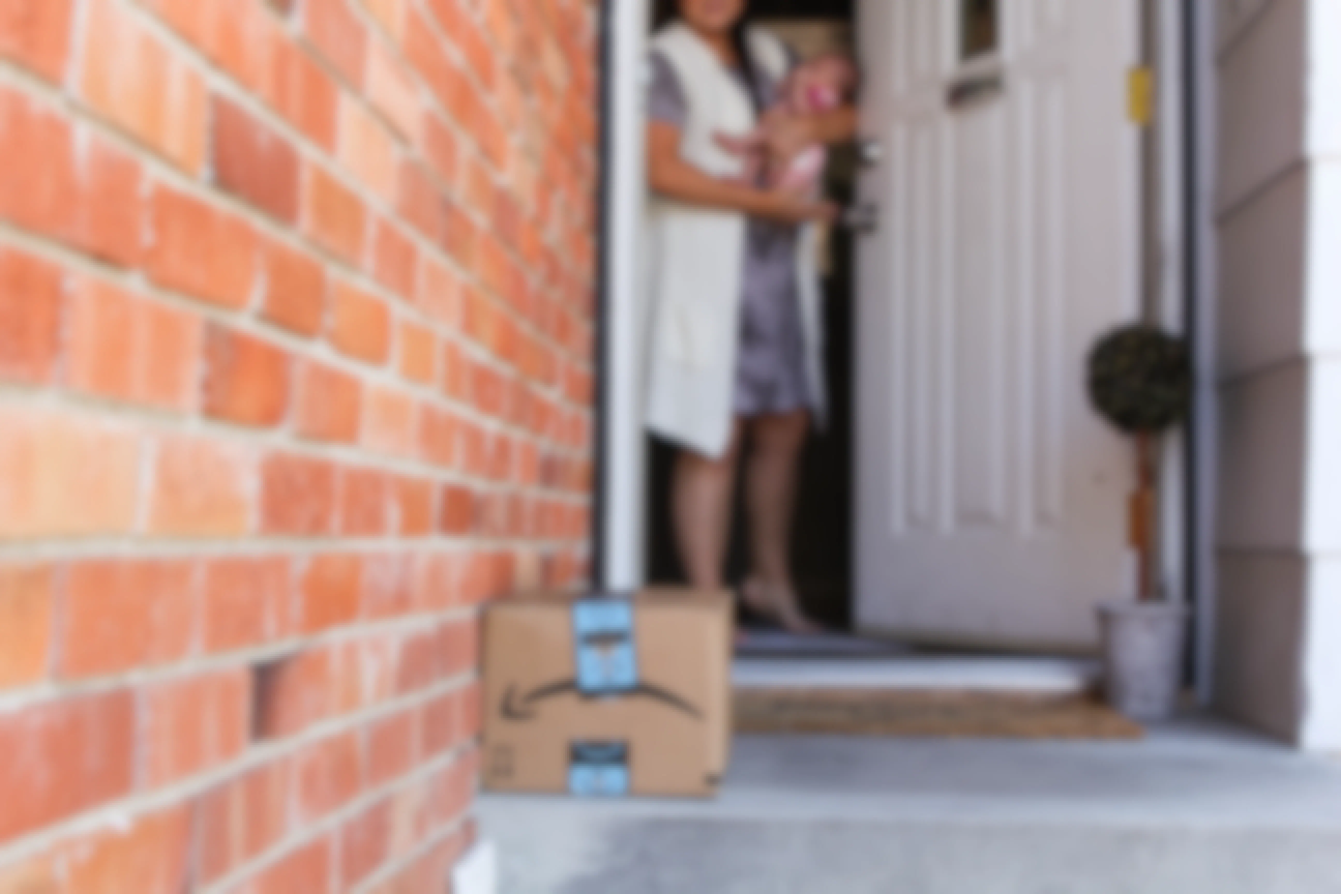 A mother holding her baby, opening the front door and looking down at an Amazon package sitting on her front porch.