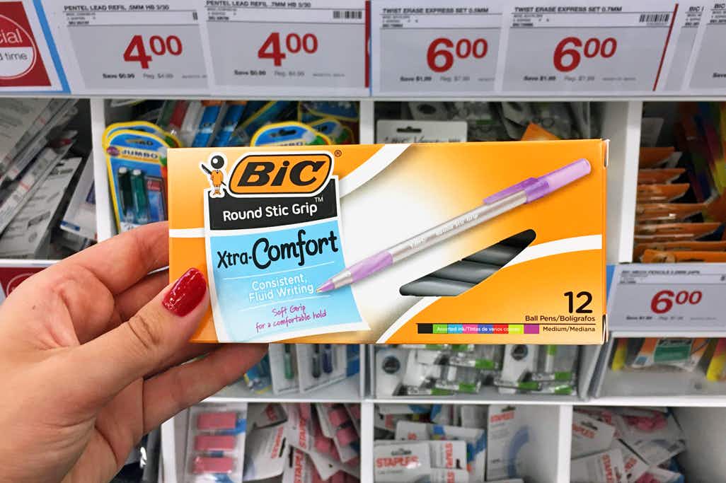 A person's hand holding a box of Xtra-Comfort Bic pens in front of a shelf inside Staples.