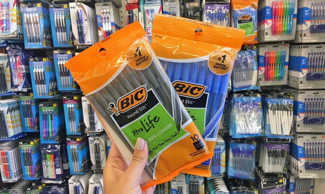 A person's hand holding two packages of BIC pens in front of a wall of pens inside Dollar Tree.