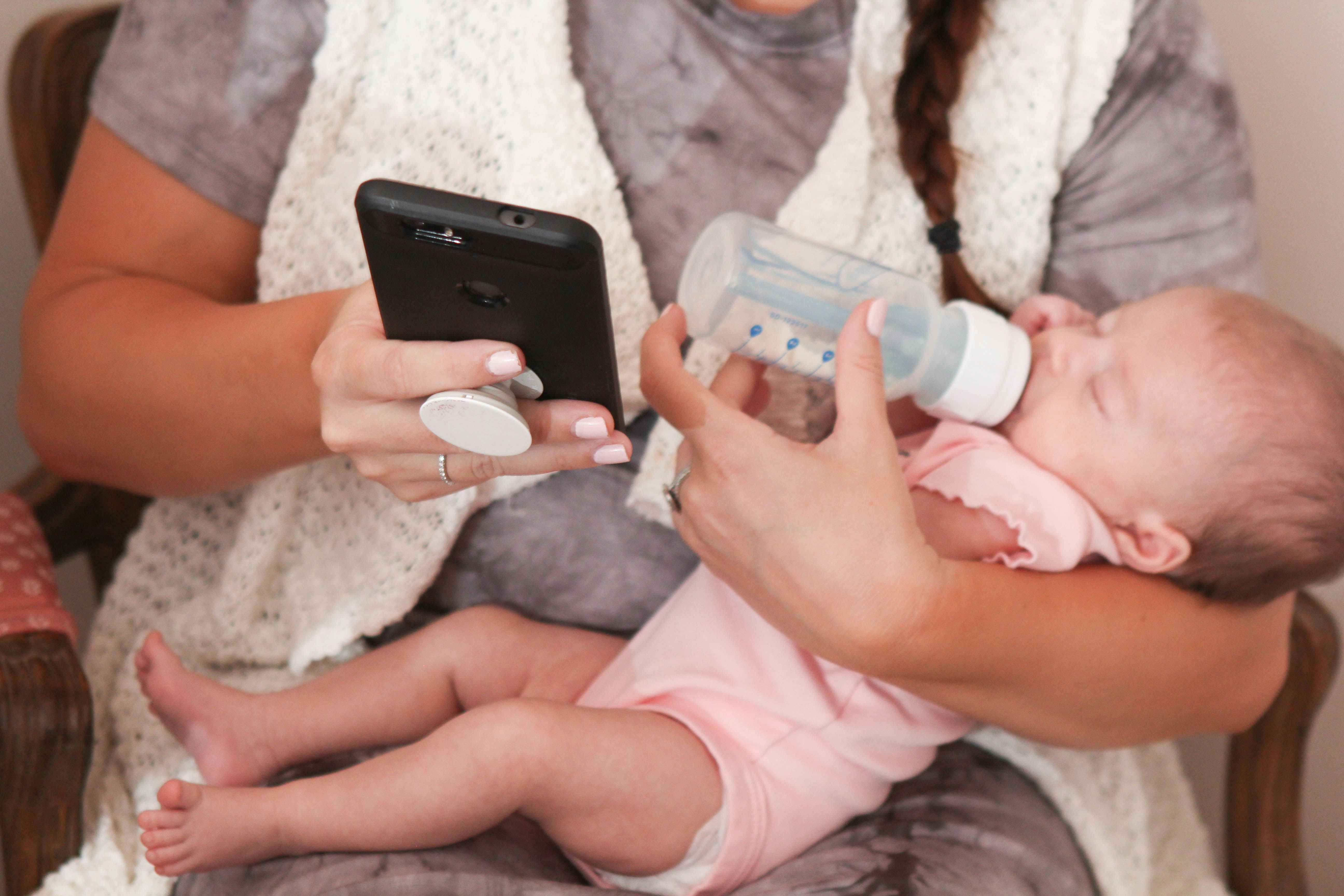 A mother sitting down and feeding a bottle to her baby while looking at her cellphone.