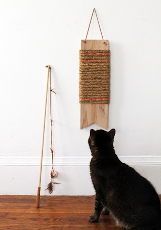 a cat sitting in front of a diy scratching pad made from a wood board and rope hung up on the wall