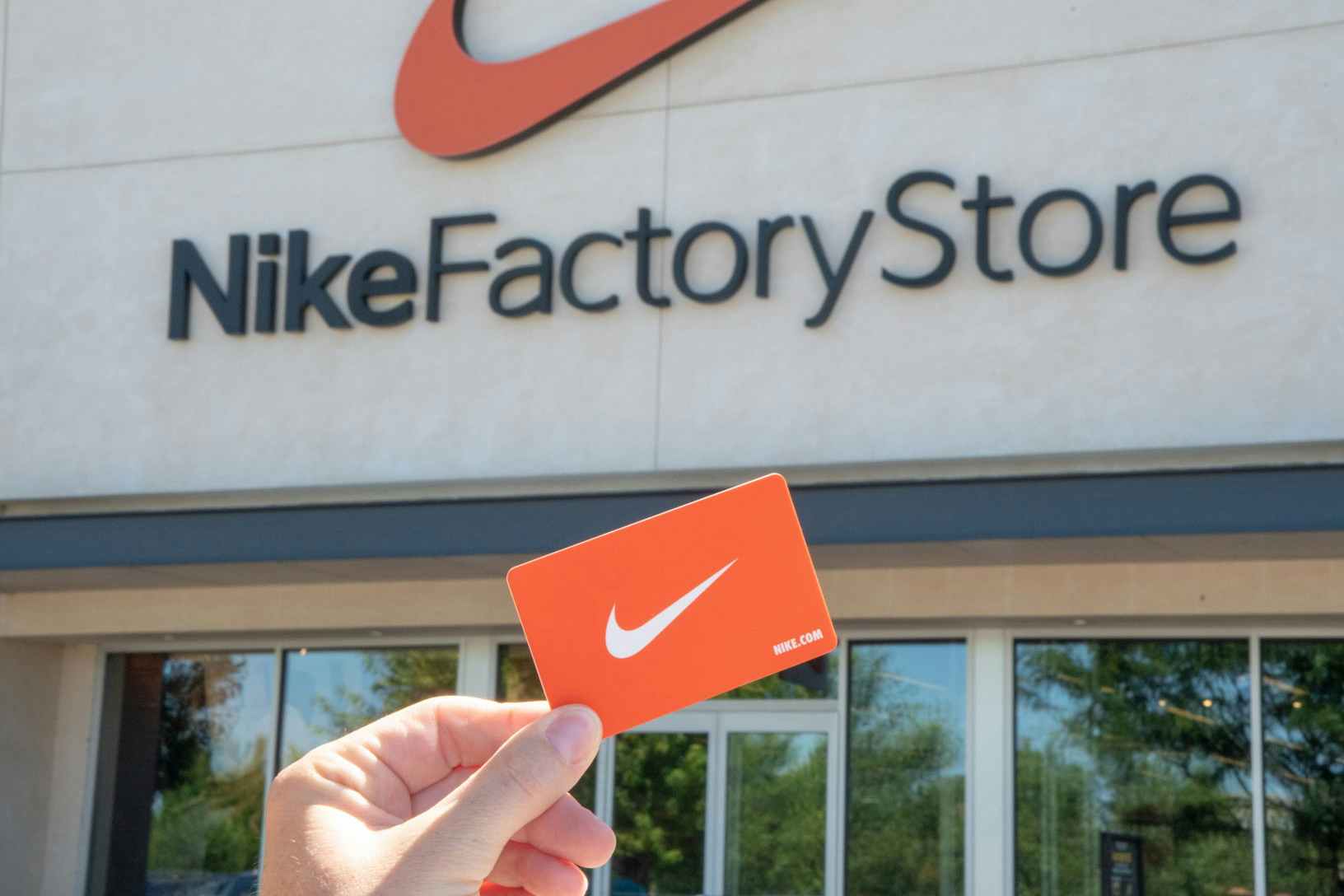 A person's hand holding up an orange Nike gift card in front of a Nike Factory Store storefront.