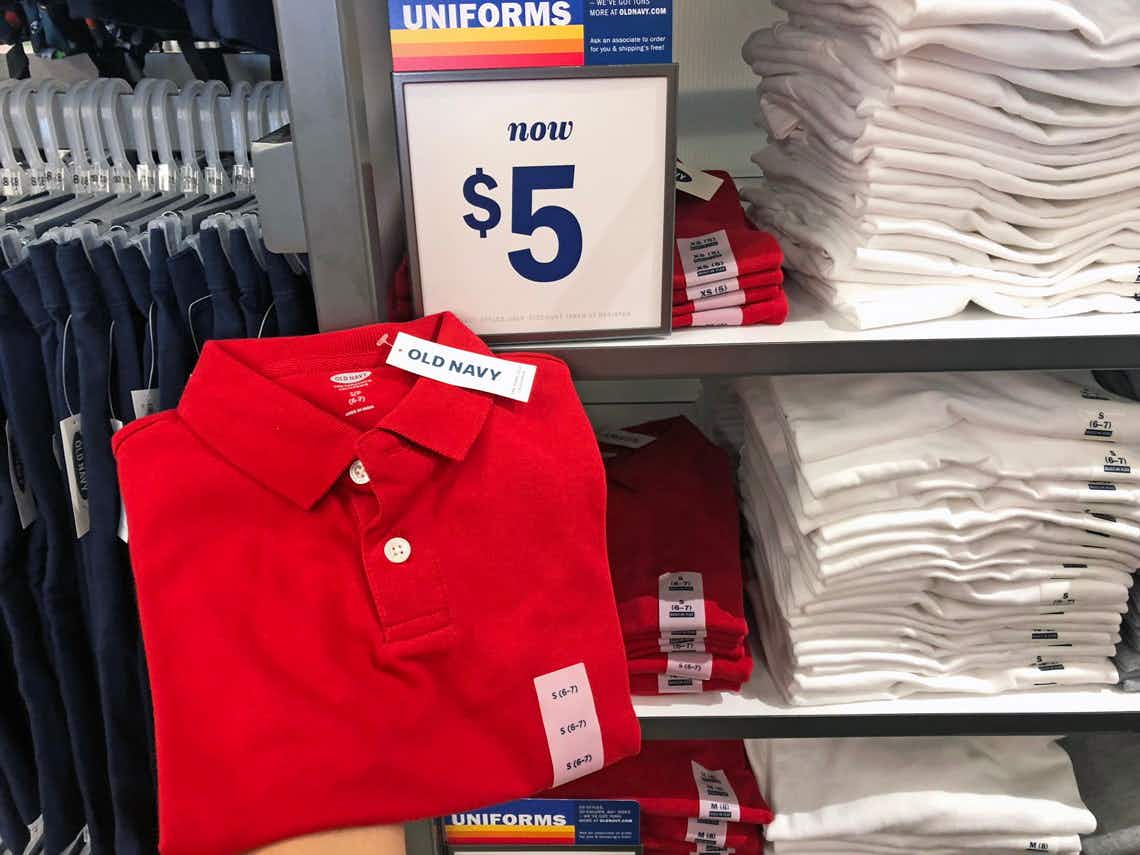 A folded kids' polo shirt being held up in front of a shelf of more kids' polo shirts and a sign that reads, "Uniforms, now $5" inside Old Navy.