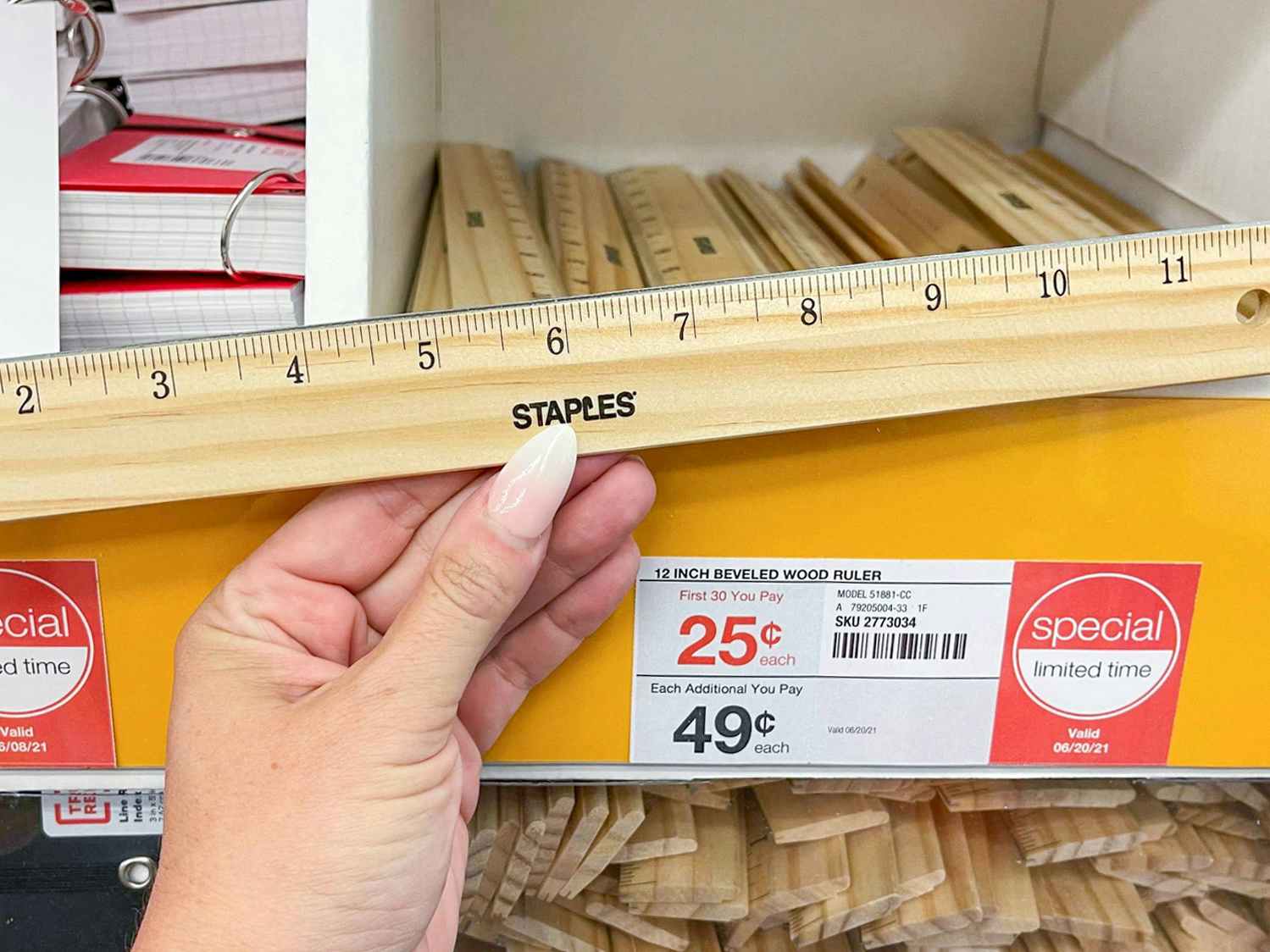 A person's hand holding a Staples ruler in front of a shelf with more rulers on sale for $0.25 at Staples.