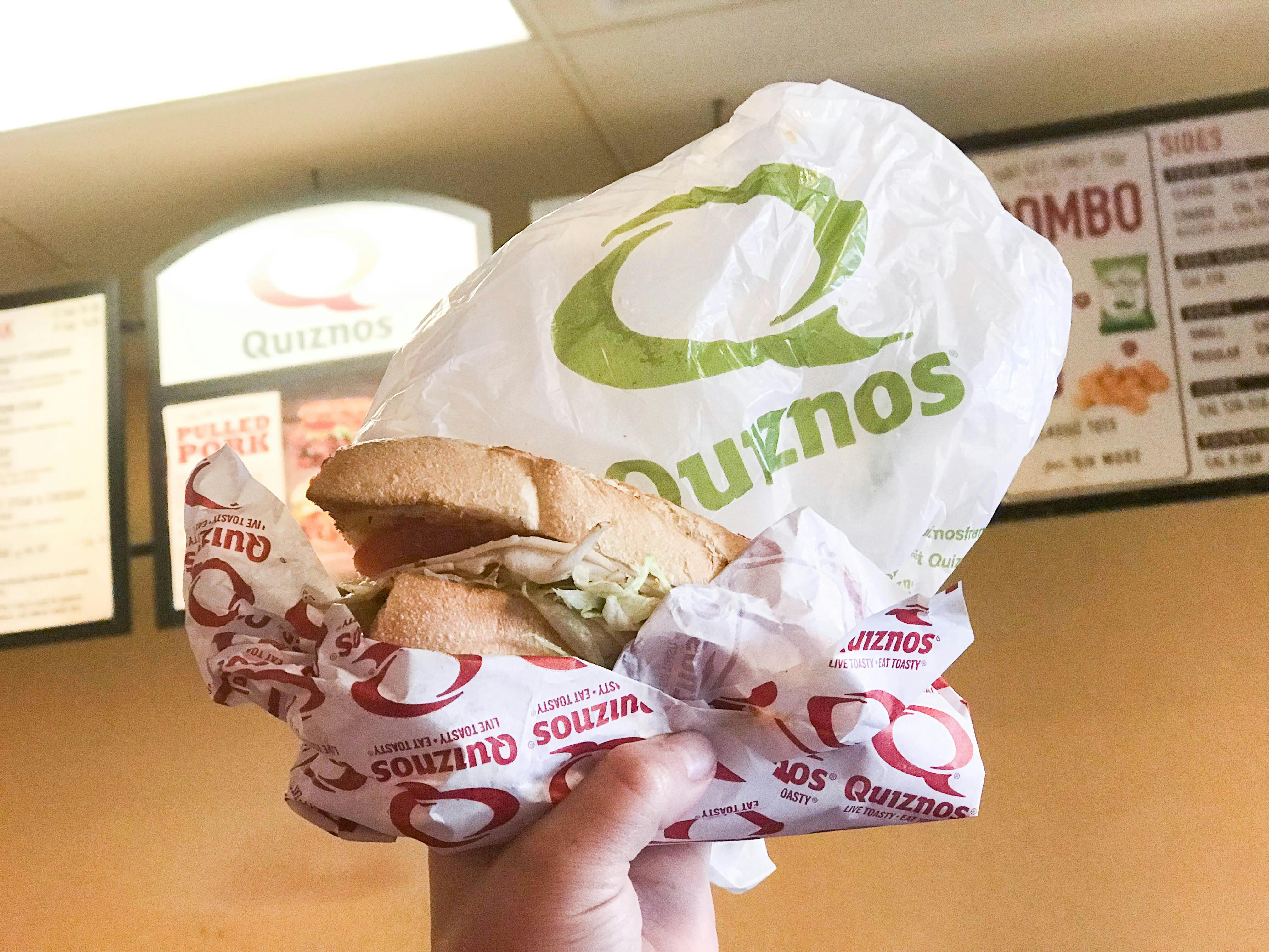 hand holding a four inch Quiznos sub in front of the Quiznos menu on National Sandwich Day