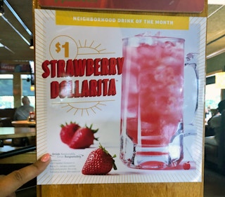 A sign for Applebee's Neighborhood Drink of the Month, advertising the $1 Strawberry Dollarita, with a person's hand pointing to the text that reads