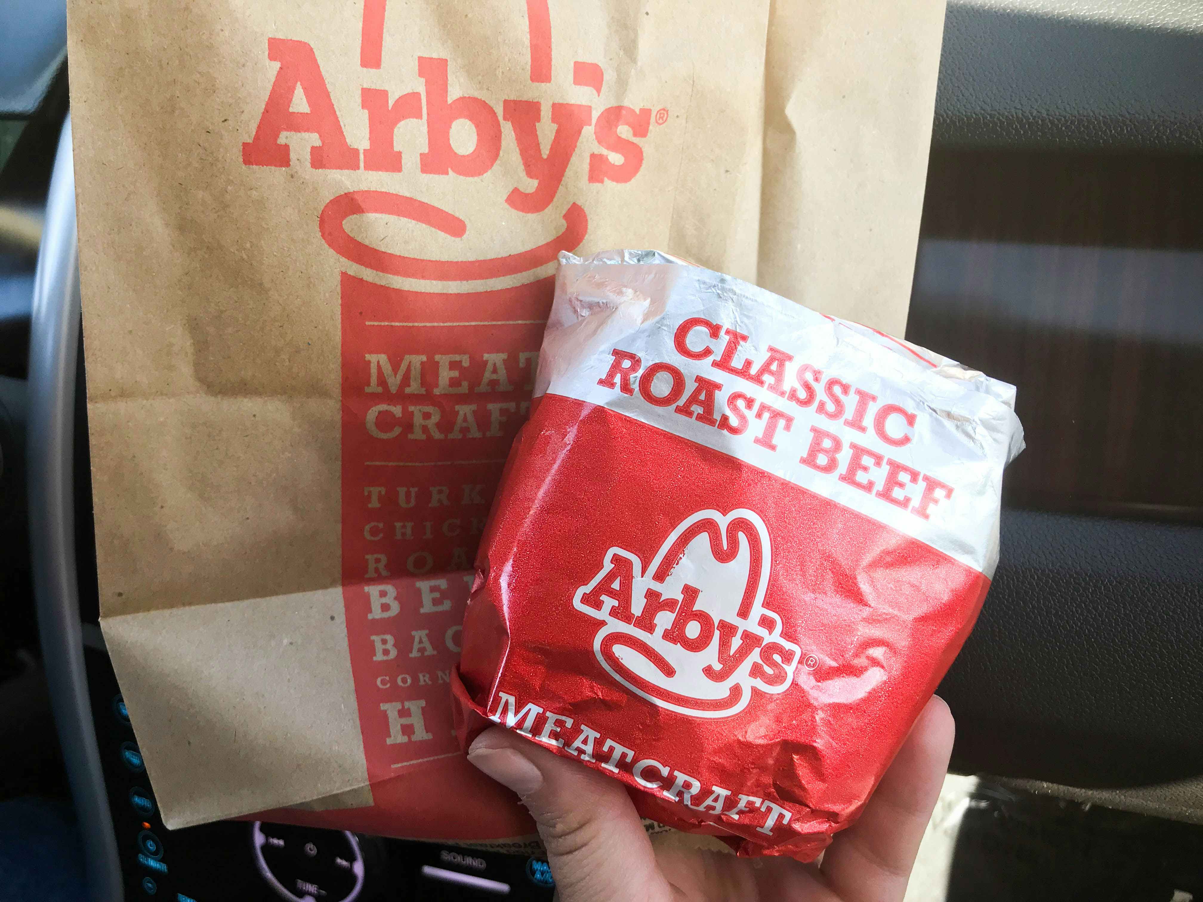 A person's hand holding an Arby's classic roast beef sandwich in its wrapper and an Arby's takeout bag in a car.