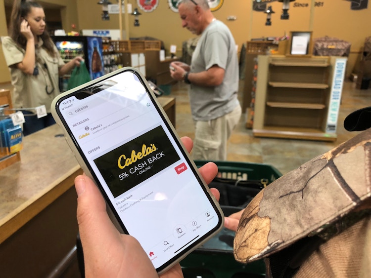 A person's hand holding their phone that is displaying the Cabela's rebate reward on the iBotta app.