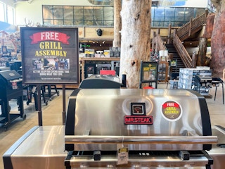 A barbecue grill on display next to a sign that reads, "Free Grill Assembly" inside Cabela's.