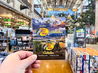 A person's hand holding a Bass Pro Shops/Cabela's gift card inside a Cabela's store.