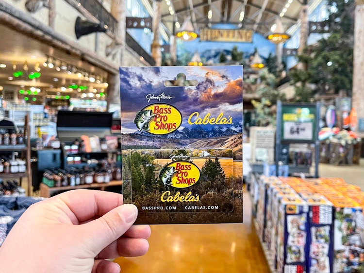 A person's hand holding a Bass Pro Shops/Cabela's gift card inside a Cabela's store.