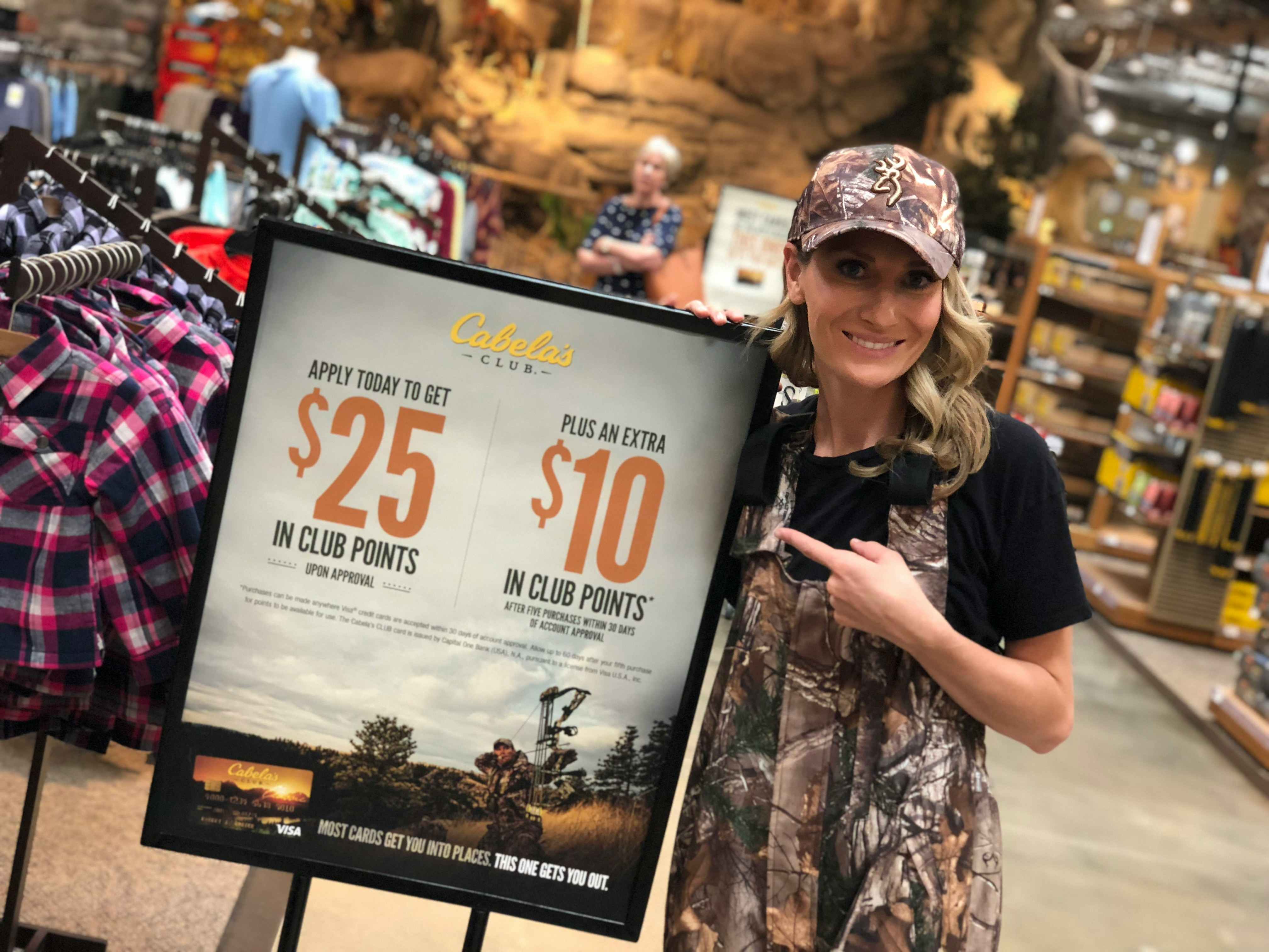 A woman standing next to and pointing at a Cabela's Club sign advertising the perks of applying for the credit card.