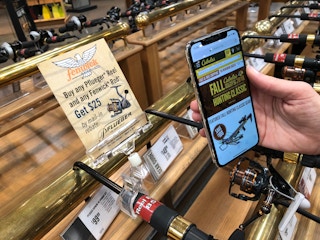 A person's hand holding their cellphone, with the Cabela's site open, in front of a display of fishing rods.